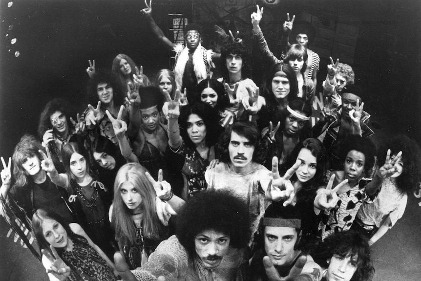 A black and white photo of the original broadway cast of hair all with their hands doing the peace sign