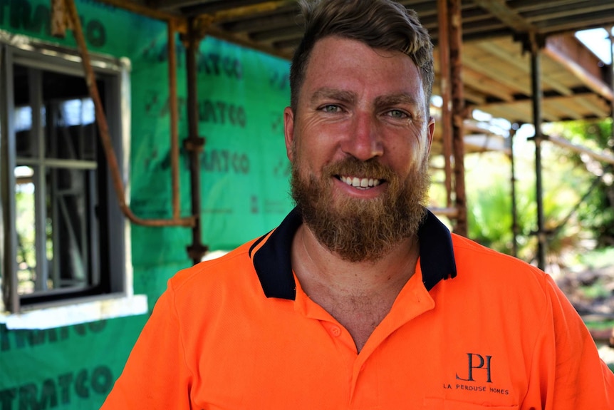 A man stands outside looking into the camera, wearing an orange shirt and smiling.