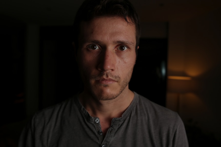 Man wearing a dark grey shirt, standing in a dark room.