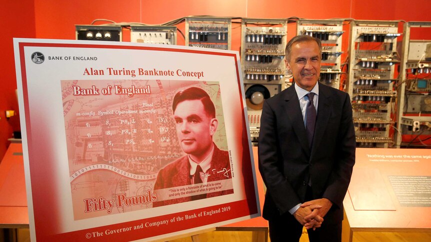 A man in a dark suit smiles in a red room in front of a large placard that reads 'Alan Turing Banknote Concept'.