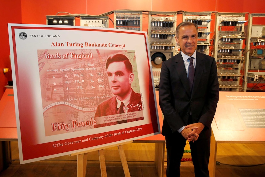 A man in a dark suit smiles in a red room in front of a large placard that reads 'Alan Turing Banknote Concept'.