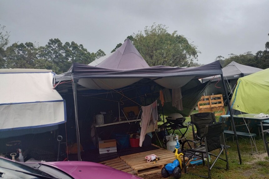 Faded purple tent, clothes hanging, black folding chair, children's toys, pink car, other tents in the background, overcast sky.