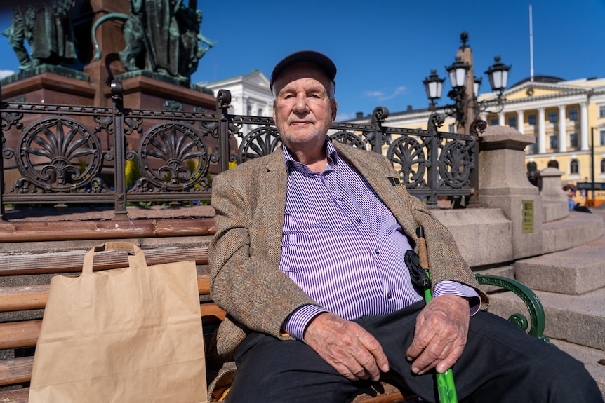 An older man in a purple shirt and brown jacket sits on a bench in front of a statue.