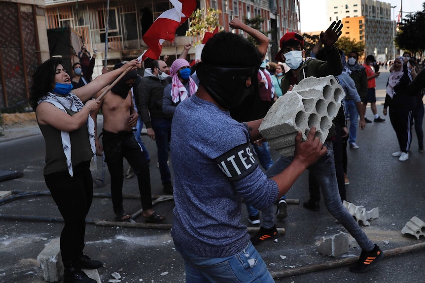 A group of young people, some with masks, wave flags and lift their arms in a shadowy city street.