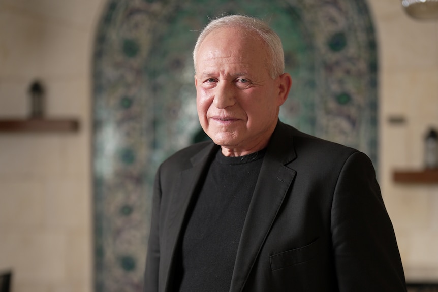 A man wearing a black suit jacket over a black shirt stands in a room looking at the camera, behind him is a tiled wall.