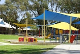 A wide shot of a school playground under blue and yellow shade sails with buildings in the background.