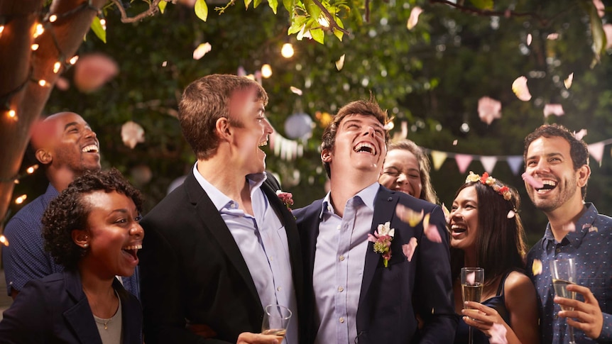 Two men hug, laugh and drink champagne on their wedding day while friends throw confetti.