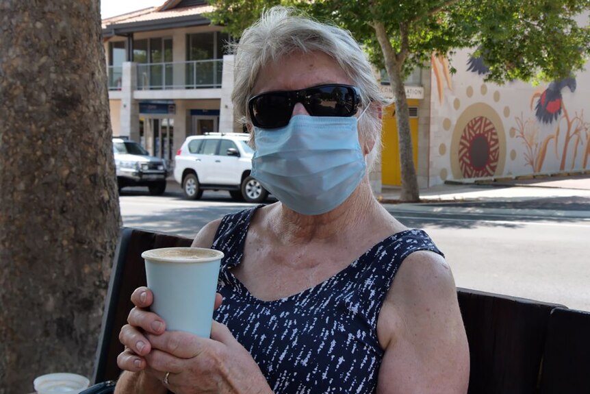 An older woman wearing a face mask and sunglasses sits on a street bench with a cup of takeaway coffee.