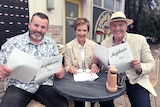Three people with Neighbours scripts seated outside a venue.