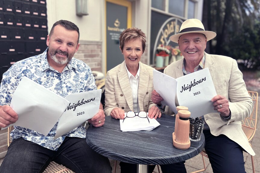 Three people with Neighbours scripts seated outside a venue.