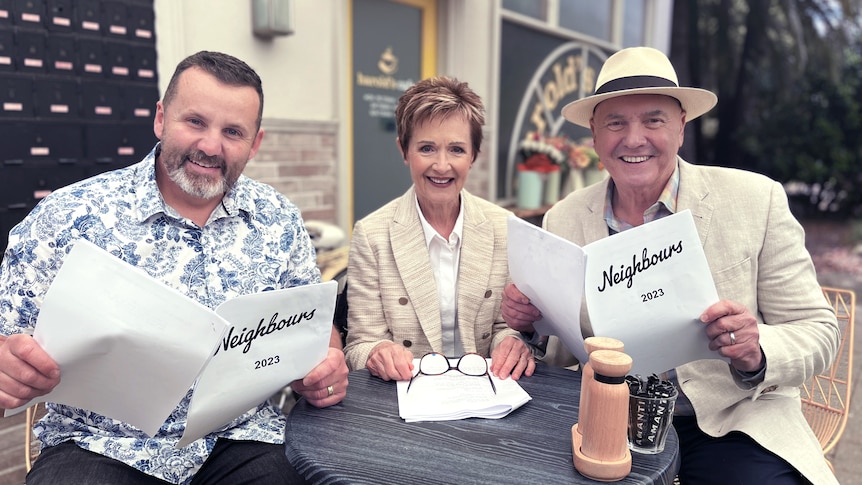 Three people with Neighbours scripts seated outside a venue.