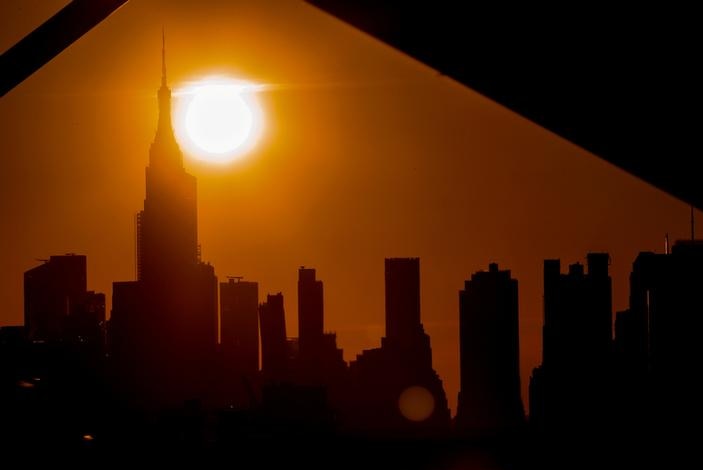 The New York City skyline is seen blacked out with the sun above the Empire State building, setting a yellow glow over it.