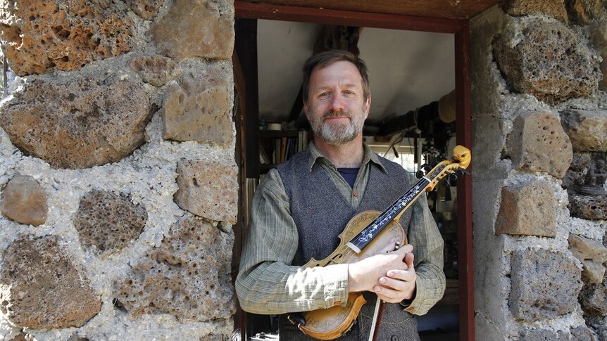 A man holding a fiddle and bow stands in the doorway of a stone-walled building.