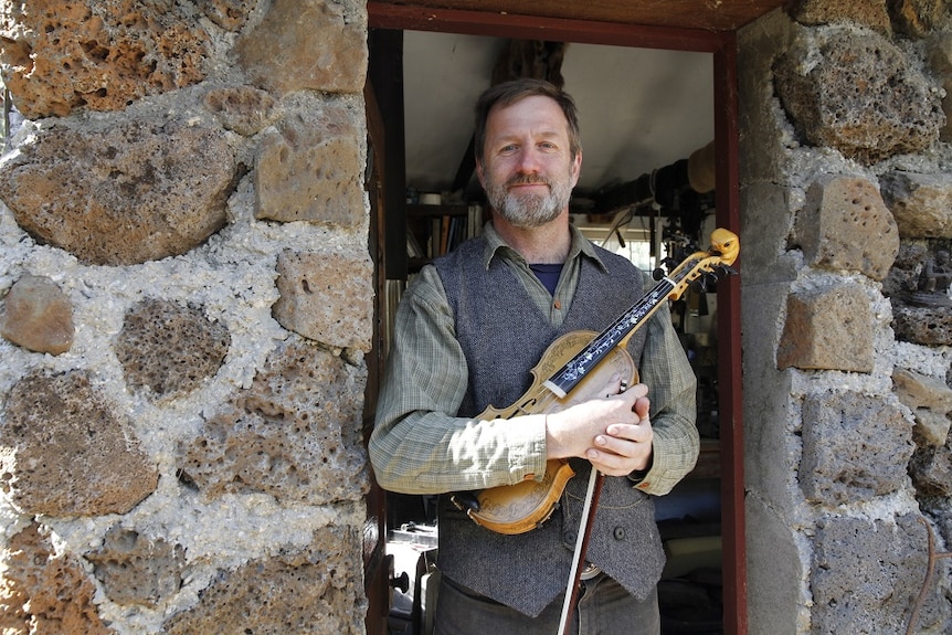 A man holding a fiddle and bow stands in the doorway of a stone-walled building.