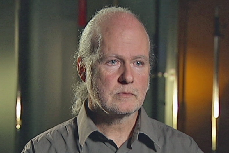 A balding man with a light-coloured beard, wearing a brown shirt, looks away at an interviewer out of shot