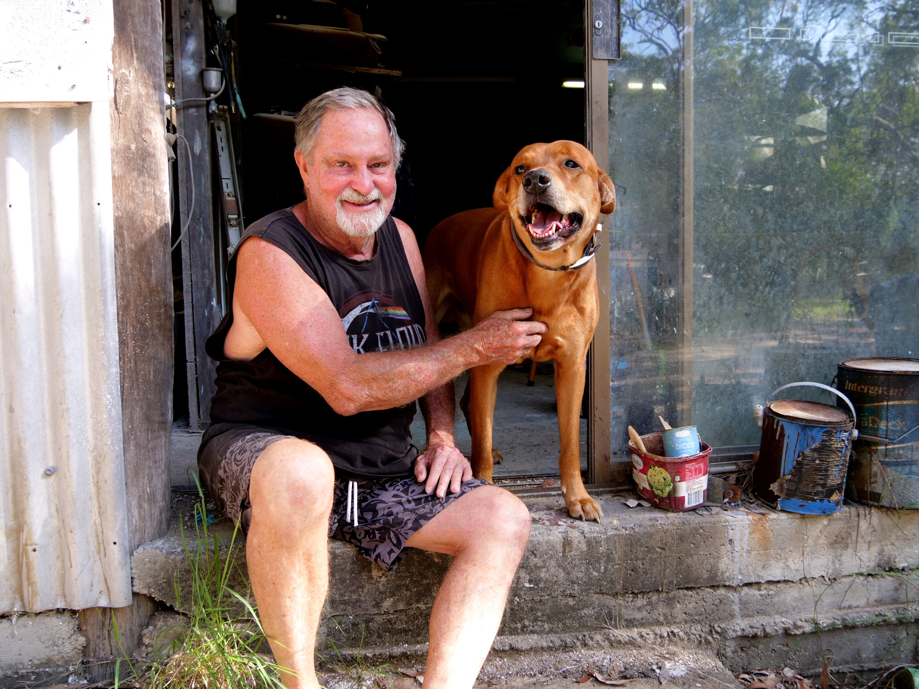 Un hombre sentado frente a un cobertizo con su perro marrón, ambos sonriendo