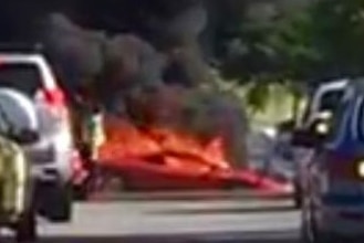 Thick black smoke rises into the sky from a burning Ferrari on a road with other cars stopped in the foreground.