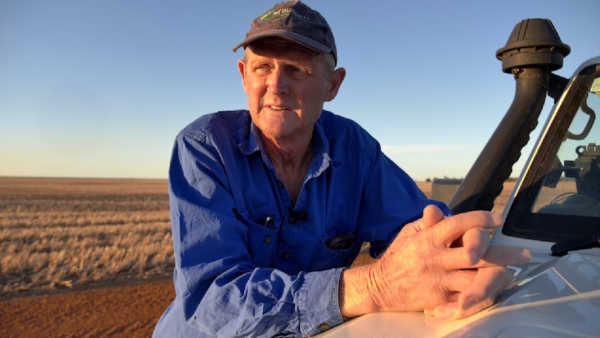 Man leaning against bonnet of a ute with hands clasped. He is wearing a blue shirt and dark cap, with fields and sky behind him