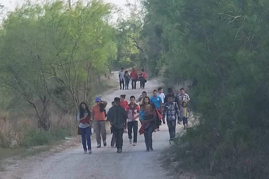 A group of about 20 men, women and children walk down a dirt path