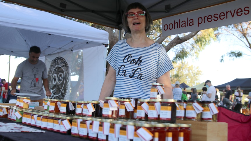 A woman stands behind a table full of jam jars at a farmers' market.