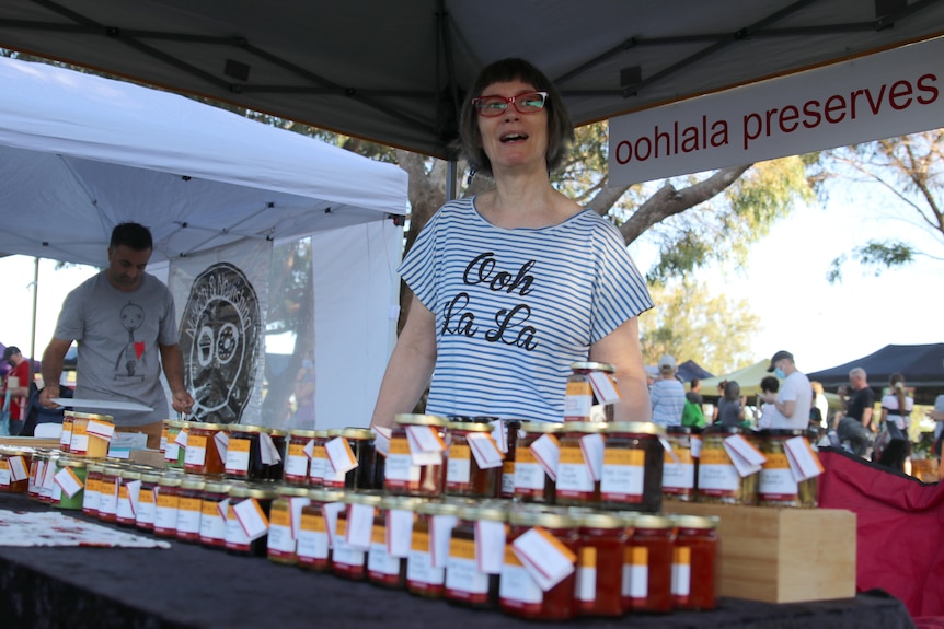 A woman stands behind a table full of jam jars at a farmers' market.