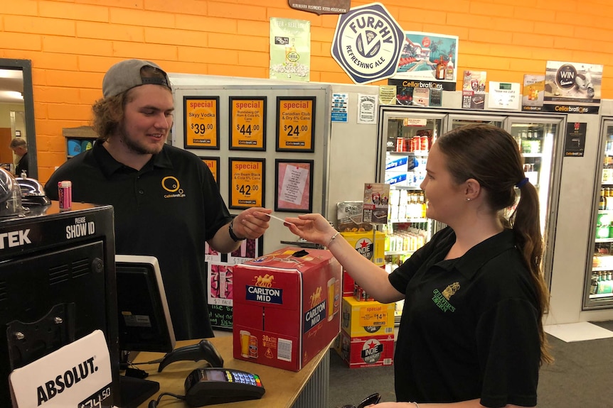 A young woman hands money to a young man behind the counter at a bottleshop