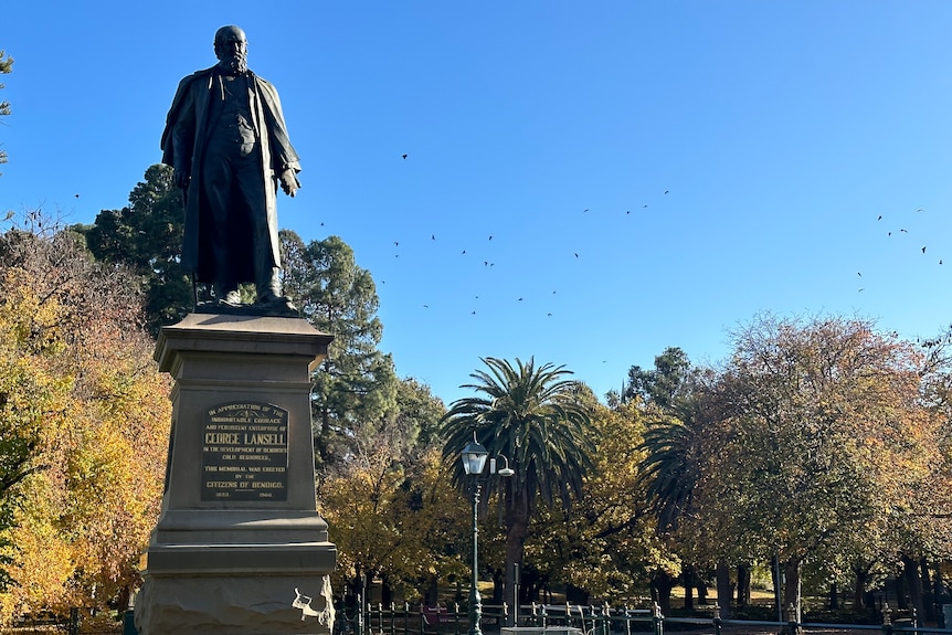 A statue of a man with bats surrounding in the sky over a nearby park.