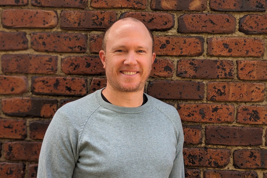 A man smiles in front of a brick wall.
