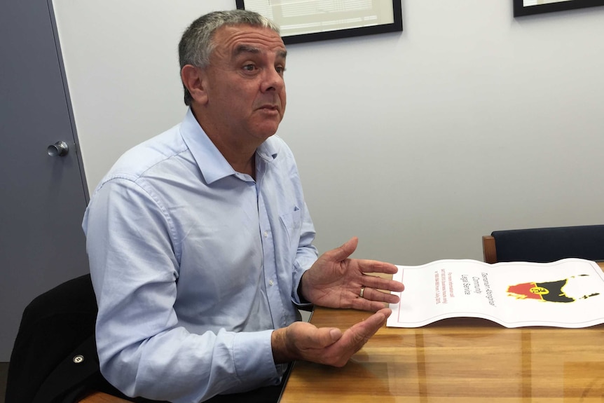 Wayne Muir wears a blue shirt and sits at a desk.