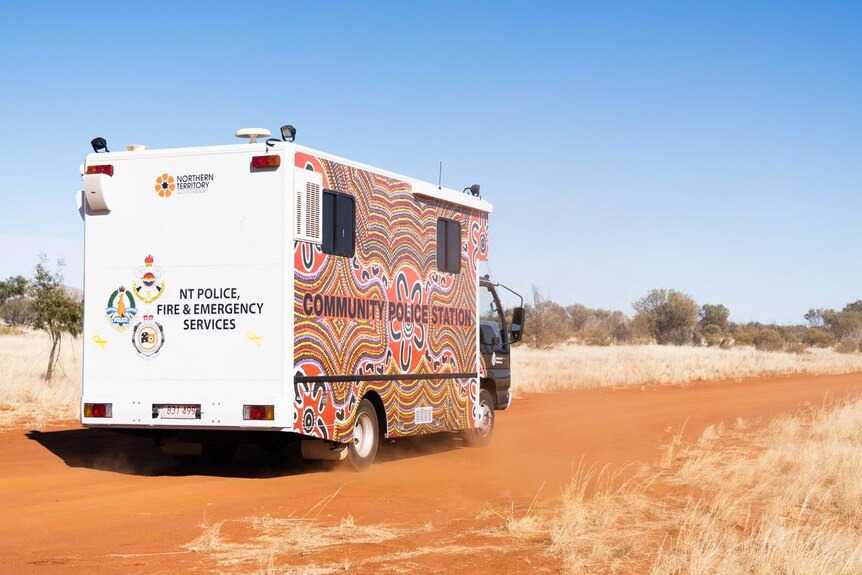 A RV-type truck covered in Indigenous art, with the words 'Community Police Station' and 'NT Police' on it, on a red dirt road.