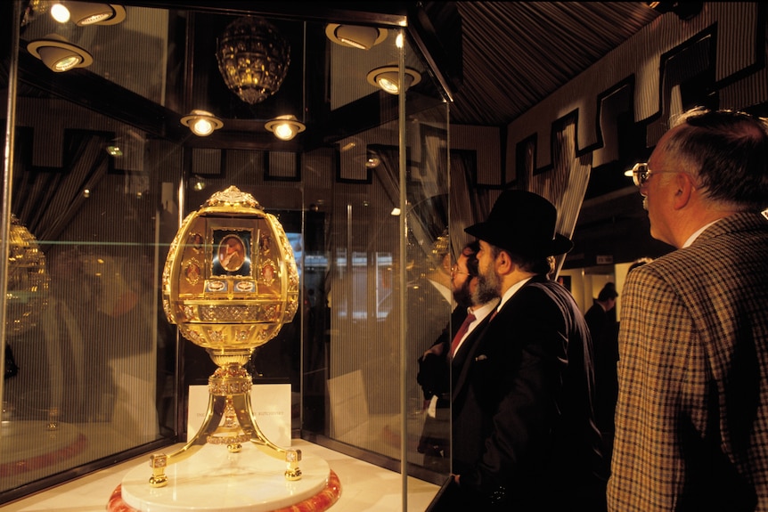 A large gold egg-shaped ornament is looked at by two men, one in a black hat.