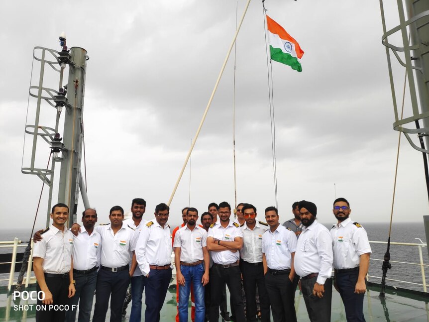 A line of ship crewmen standing on a ship with an Indian flag flying behind them and the sea in the background