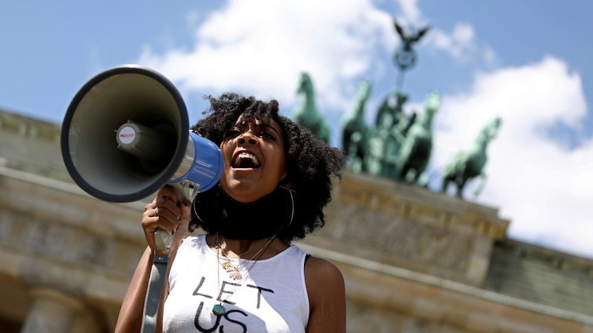 A woman holds a megaphone while wearing a white singlet that reads 'let us breathe', in front of the Brandenburg Gate in Berlin