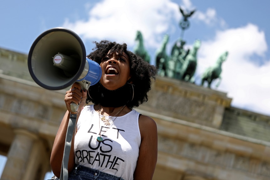 A woman holds a megaphone while wearing a white singlet that reads 'let us breathe', in front of the Brandenburg Gate in Berlin