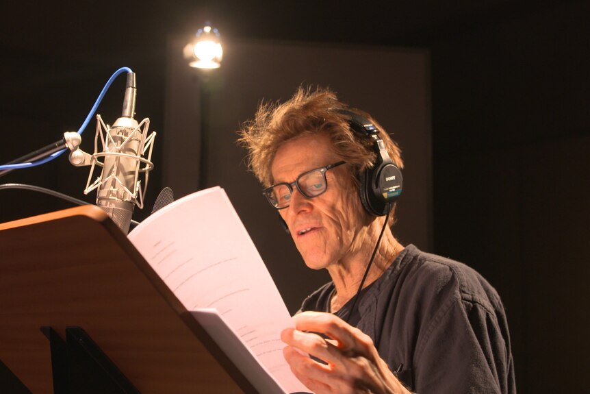 White man with weathered face, soft brown hair and glasses wears black shirt and reads from script in audio recording booth.