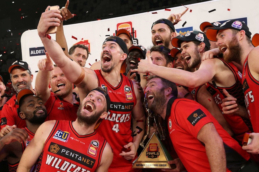 Perth Wildcats forward Greg Hire takes a team selfie with the NBL championship trophy has his teammates celebrate.