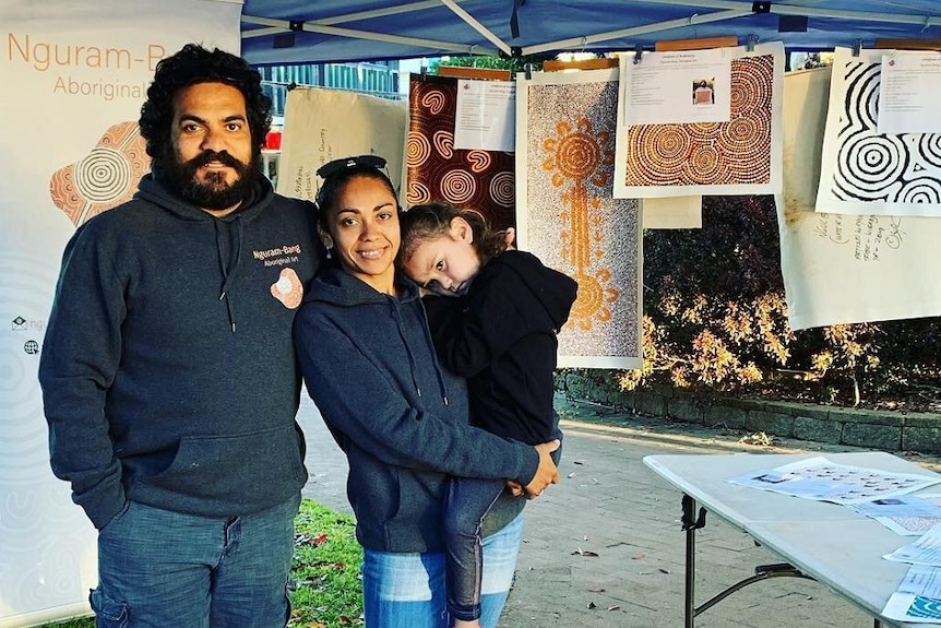 Wayne Martin and Dani Beezley holding their daughter Stella-May, standing under marquee, Indigenous artworks hanging around.