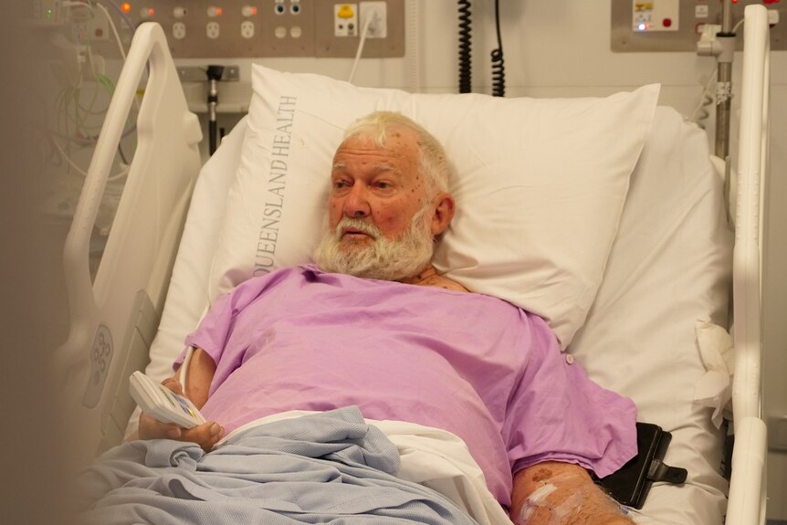 John an older man with white beard and hair lying in a hospital bed, purple gown.