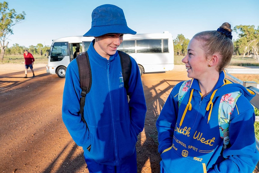 Two students in tracksuits stand next to their farm gate in front of a bus