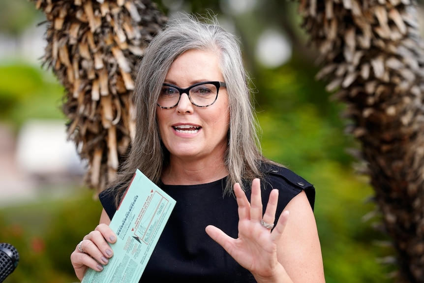 Katie Hobbs, a woman with straight grey hair and black-rimmed glasses, holds a ballot paper in her hand