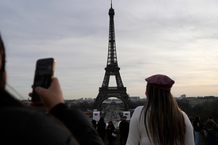 Tourists look at the Eiffel Tower from afar. One girl is wearing a red beret and another is holding up an iphone.