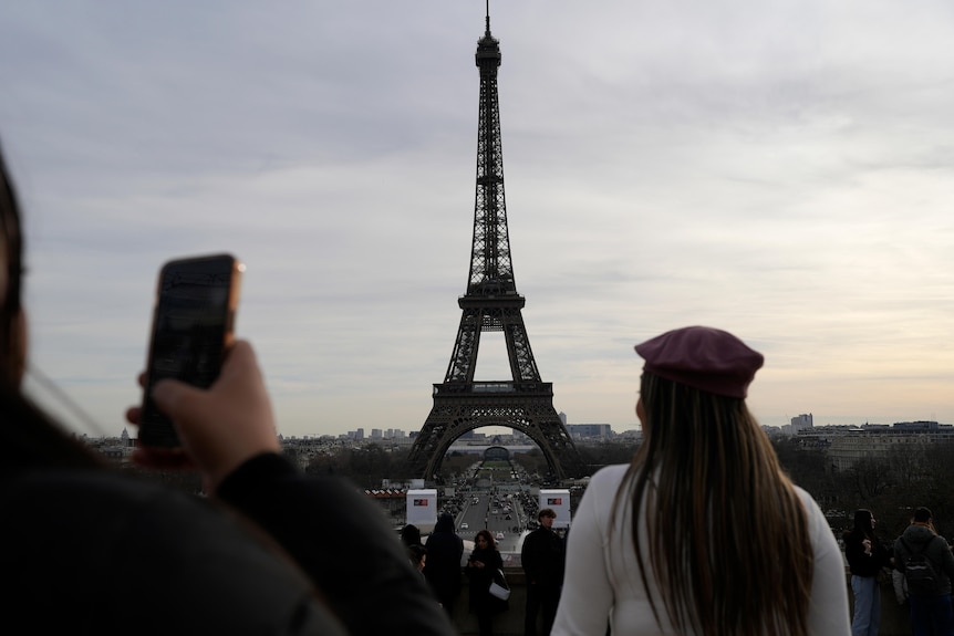 Туристы смотрят на Эйфелеву башню издалека.  Одна девушка в красной шляпе, а другая держит в руках iPhone.
