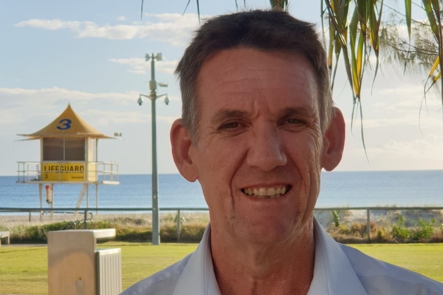 A man smiles at the camera, with a lifeguard lookout and beach behind him on a sunny day. 