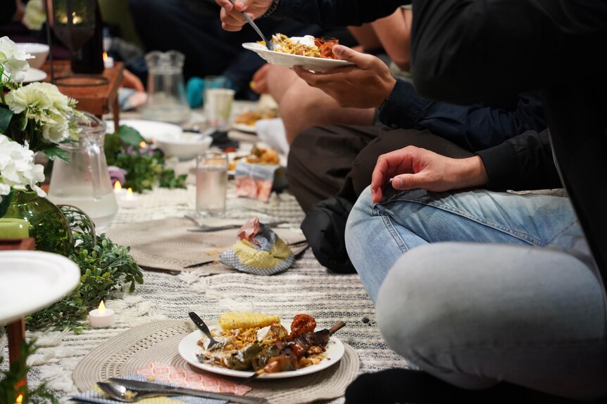 A row of people from the waist down sitting on pillows and carpet on the floor eating food from plates.