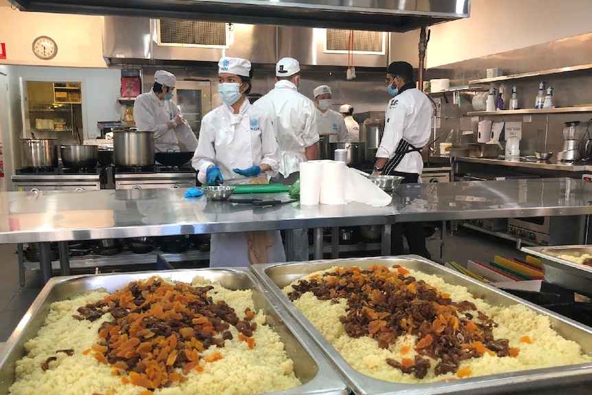 Six chefs in white kitchen clothes work at benches in a commercial kitchen.