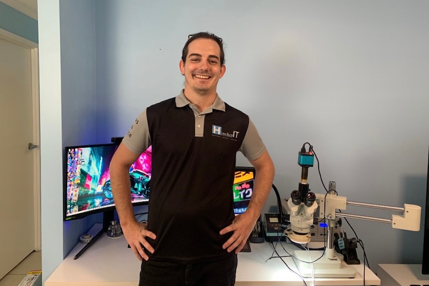 A white man with brown hair stands and smiles in front of a desk, on which sits computer screens and electronic repair equipment