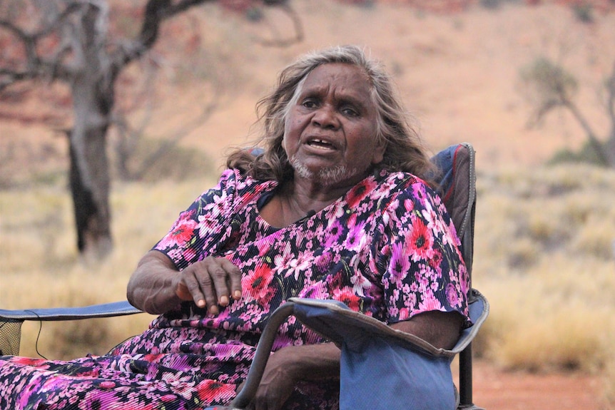 She sits in a camp chair and wears a black skirt and shirt with purple and red flowers