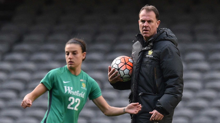 Alen Stajcic stands hugging a soccer ball to his chest while watching a Matildas player during a training session.