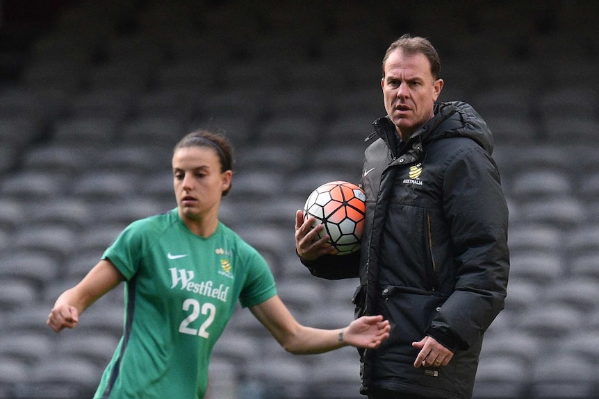 Alen Stajcic stands hugging a soccer ball to his chest while watching a Matildas player during a training session.