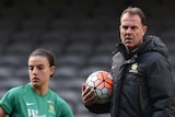 Alen Stajcic stands hugging a soccer ball to his chest while watching a Matildas player during a training session.